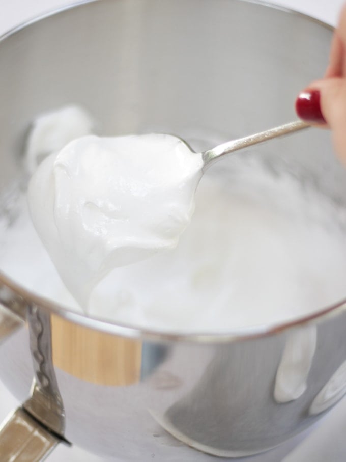 Scoop of uncooked meringue in kitchen aid mixer bowl on a spoon with red fingernail for recipe on how to make a pavlova.