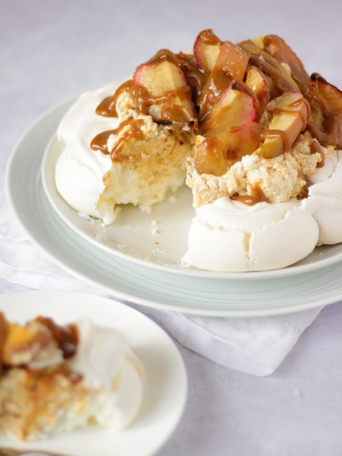 Salted caramel and apple pavlova on a white plate on white background piled with apples with slice cut out on a plate in the foreground.