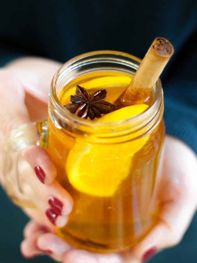 Person in a green jumper with red painted nails holding a glass mug of slow cooker mulled cider face and legs not visible. With orange, cinnamon stick and star anise in glass.