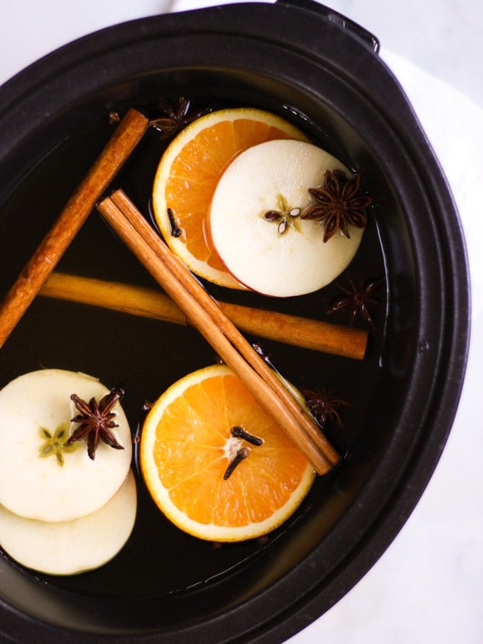 Overhead photo of ingredients in a slow cooker for mulled cider, including cider (cyder) cut orange, apple, cinnamon sticks, star anise, cloves.