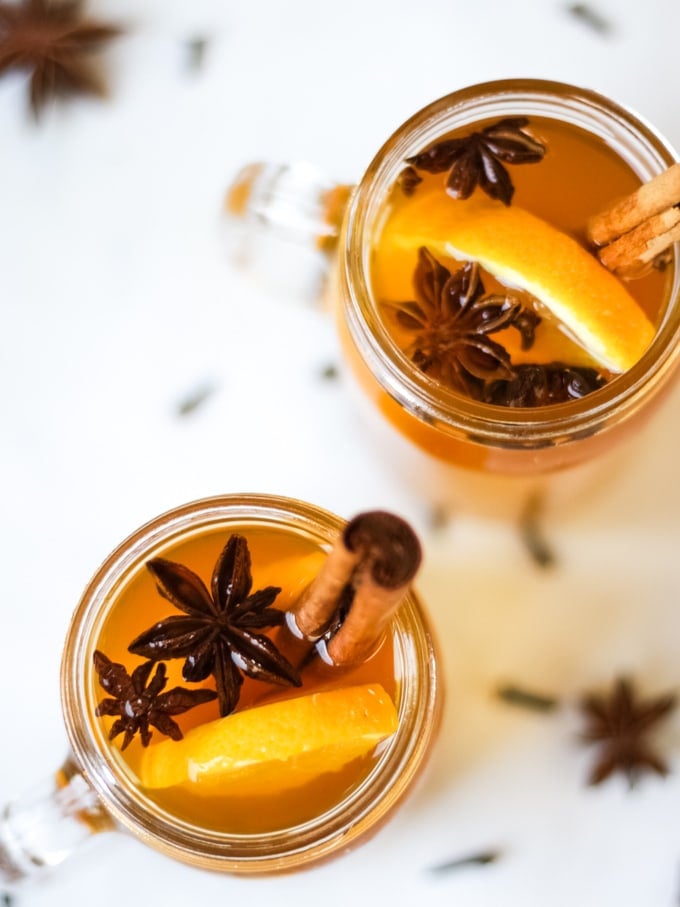 Overhead photo of two glass mugs of slow cooker mulled cider recipe on a white background, with slices of orange, cinnamon sticks, star anise and cloves.