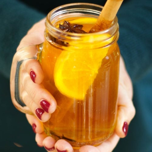 Person in a green jumper with red painted nails holding a glass mug of slow cooker mulled cider face and legs not visible. With orange, cinnamon stick and star anise in glass.
