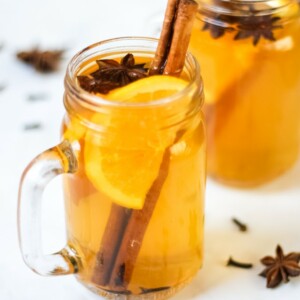 Overhead photo of two glass mugs of slow cooker mulled cider recipe on a white background, with slices of orange, cinnamon sticks, star anise and cloves.