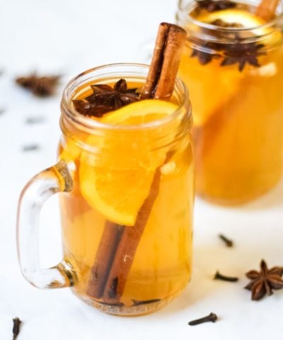 Overhead photo of two glass mugs of slow cooker mulled cider recipe on a white background, with slices of orange, cinnamon sticks, star anise and cloves.