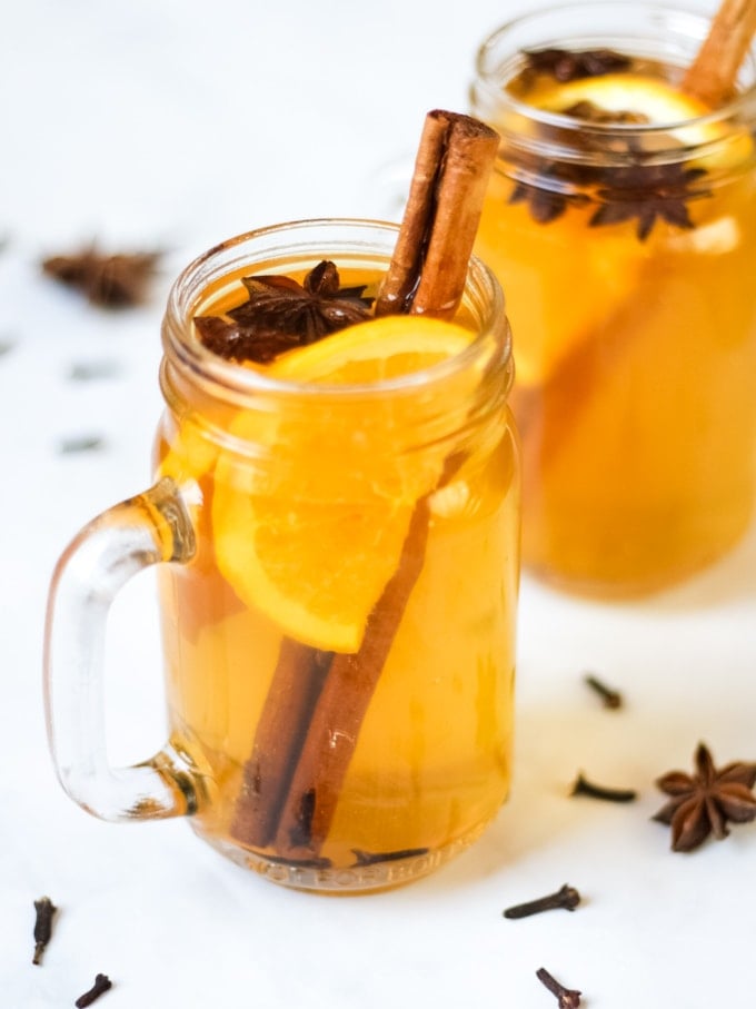 Overhead photo of two glass mugs of slow cooker mulled cider recipe on a white background, with slices of orange, cinnamon sticks, star anise and cloves.