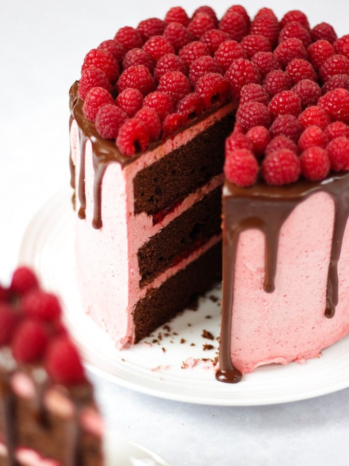 Raspberry cake on white plate on white marble background with chocolate ganache and fresh raspberries on top with wedge cut out and on a plate in the foreground.