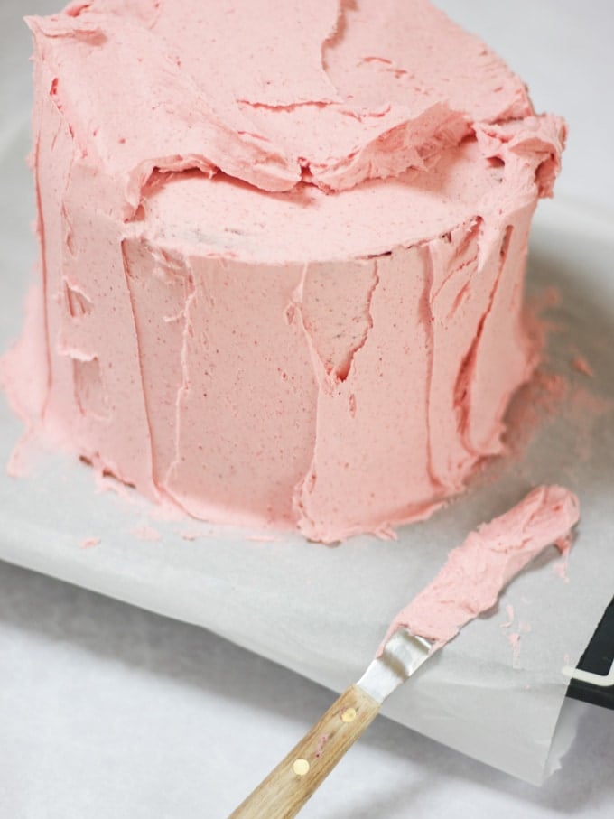 Raspberry cake receiving a second coat of buttercream frosting icing with a palette knife on baking paper.