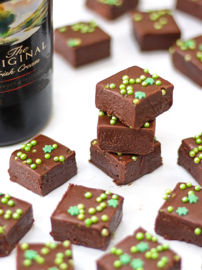 Pile of 3 pieces of easy baileys fudge and other pieces on white background with green sprinkles and bottle of baileys.