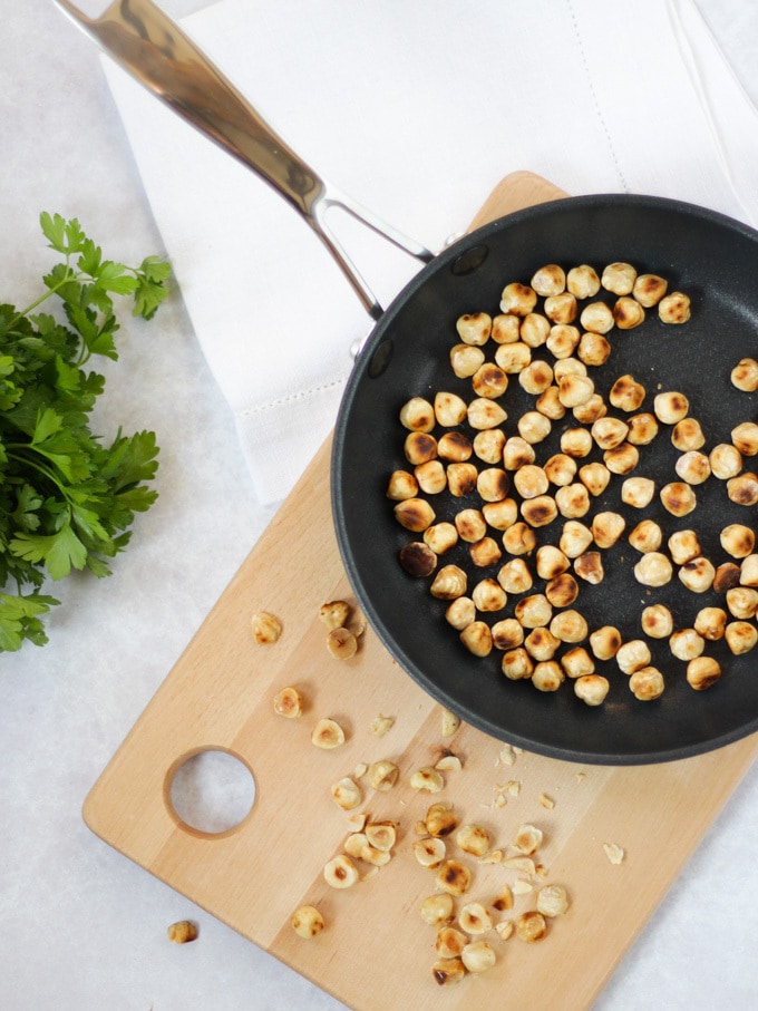 Overhead photo of black frying pan with toasted hazelnuts and wooden board and chopped herbs.