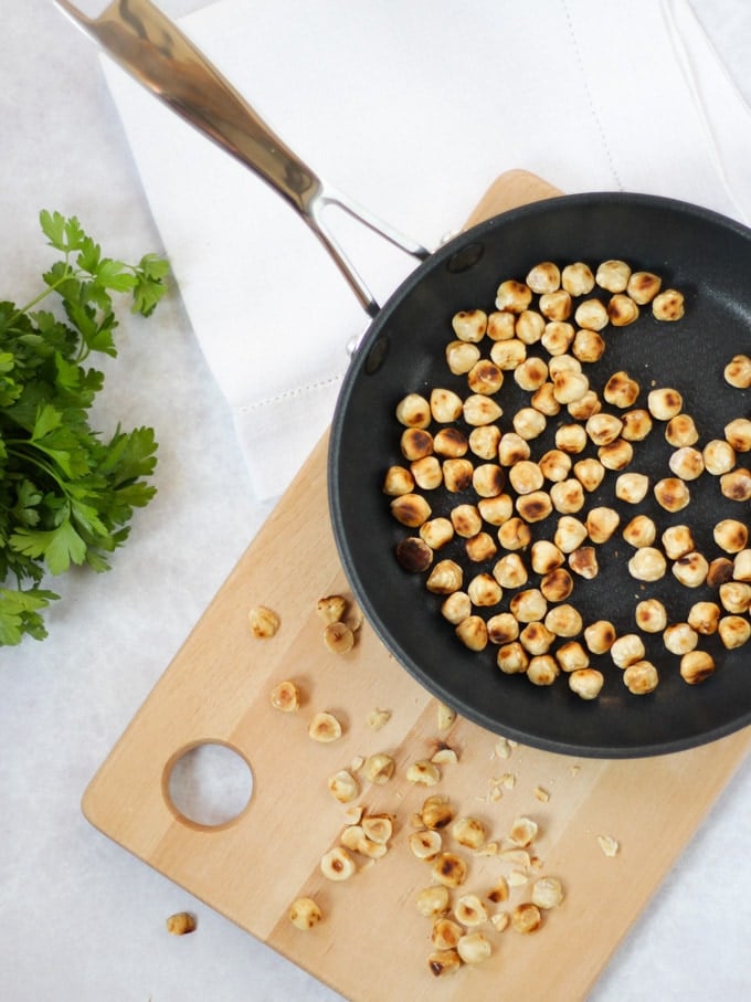 Overhead photo of black frying pan with toasted hazelnuts and wooden board and chopped herbs.