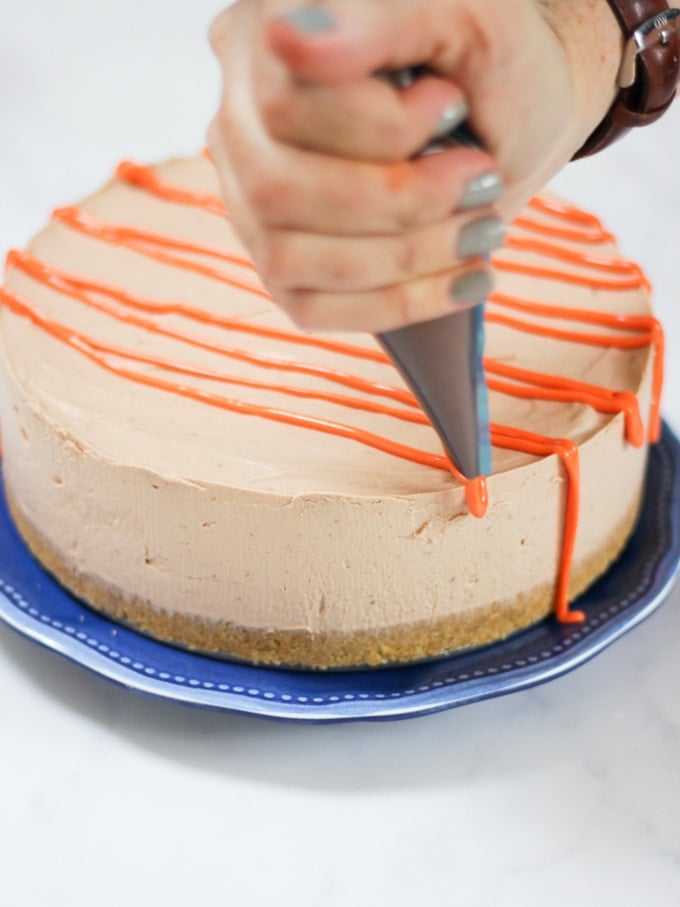 Photo of a no bake chocolate orange cheesecake on a blue plate being covered with orange chocolate drizzle from a piping bag.