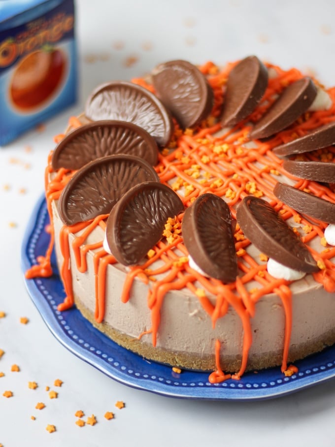 Side angle photo of a No Bake Terry's Chocolate Orange Cheesecake on a blue plate with a chocolate orange in a box in the background, drizzled with white chocolate coloured orange for this dessert recipe.