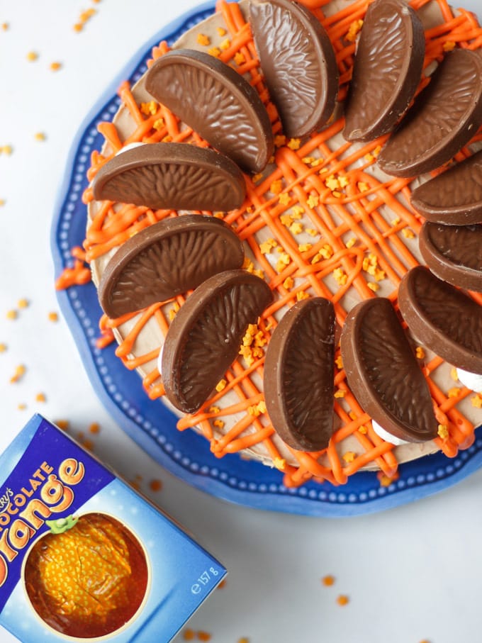 Overhead photo of a No Bake Terry's Chocolate Orange Cheesecake on a blue plate with a chocolate orange in a box in the background, drizzled with white chocolate coloured orange for this dessert recipe.