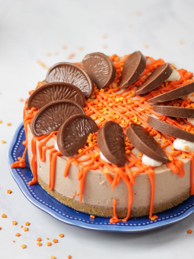 Side angle photo of a No Bake Terry's Chocolate Orange Cheesecake on a blue plate with a chocolate orange in a box in the background, drizzled with white chocolate coloured orange for this dessert recipe.
