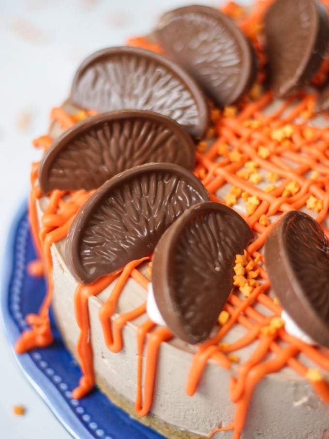 No Bake Terry's Chocolate Orange Cheesecake on a blue plate with a chocolate orange in a box in the background, drizzled with white chocolate coloured orange for this dessert recipe.