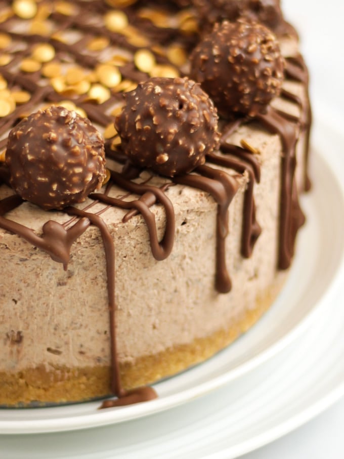 Side angle close up of a no bake Ferrero richer cheesecake recipe on a white plate with Ferrero richer and gold sprinkles on top with dropped chocolate.