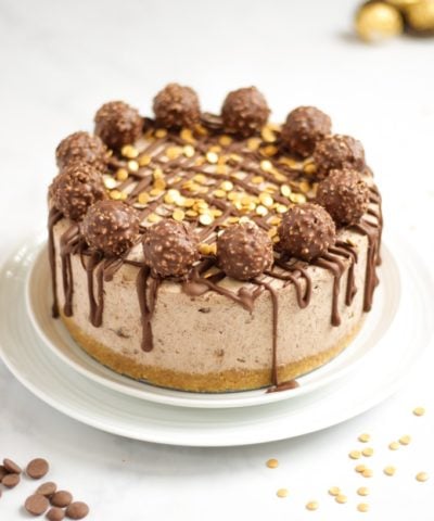 Side angle photo of a no bake Ferrero richer cheesecake recipe on a white plate with Ferrero richer and gold sprinkles on top with dropped chocolate.