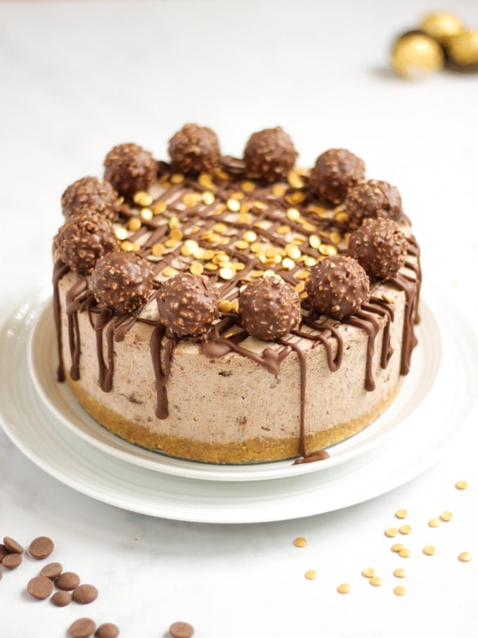 Side angle photo of a no bake Ferrero richer cheesecake recipe on a white plate with Ferrero richer and gold sprinkles on top with dropped chocolate.