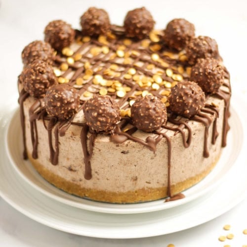 Side angle photo of a no bake Ferrero richer cheesecake recipe on a white plate with Ferrero richer and gold sprinkles on top with dropped chocolate.