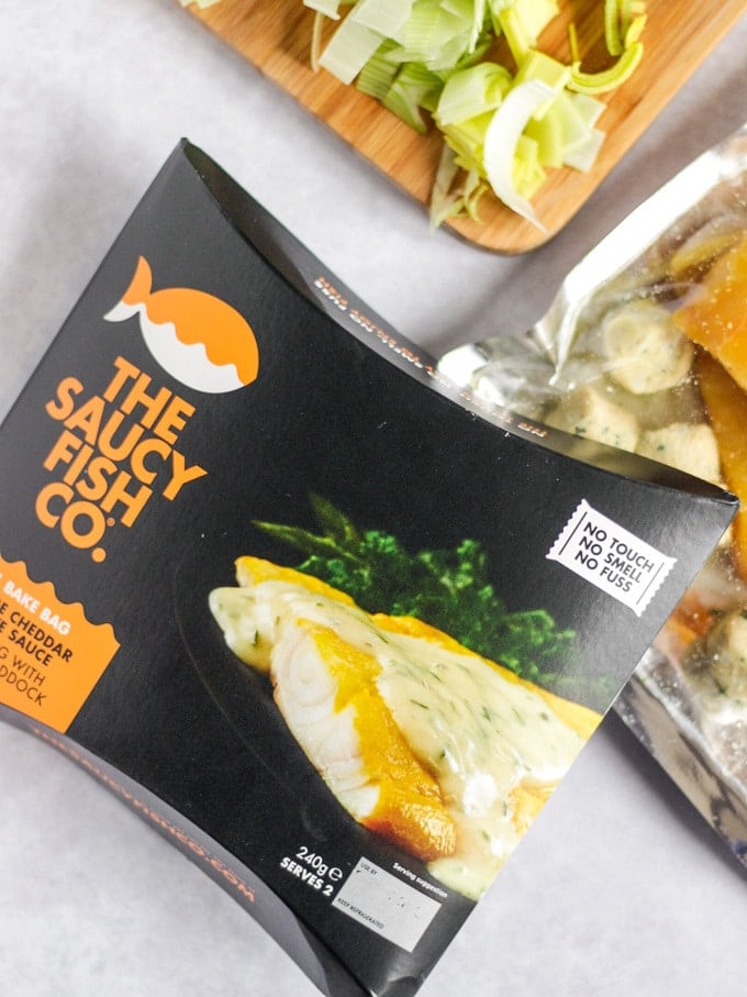 The Saucy Fish Co packet on marble background with chopped leeks on wooden board.