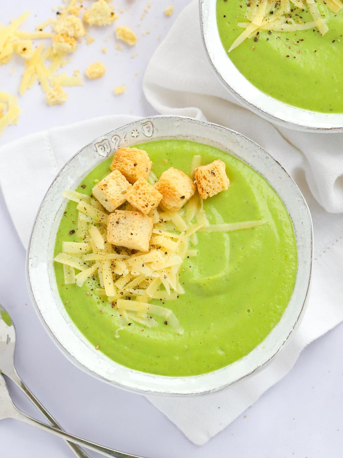 A white tablecloth with bowl filled with green Broccoli and Cauliflower Soup, topped with cheese and croutons.
