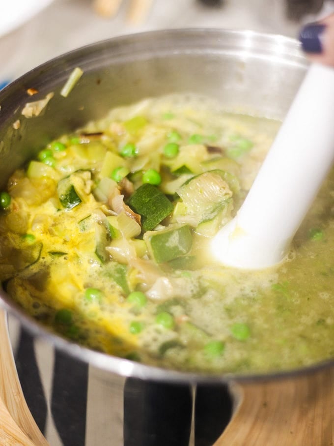 Silver pan of chopped and fried courgettes, onions, peas and stock with stick hand blender in ready to blend to courgette soup recipe.