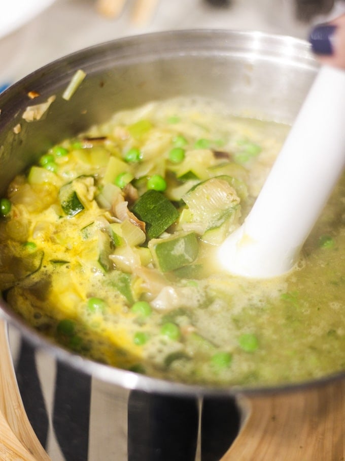 Silver pan of chopped and fried onions, peas and stock with stick hand blender in ready to blend.
