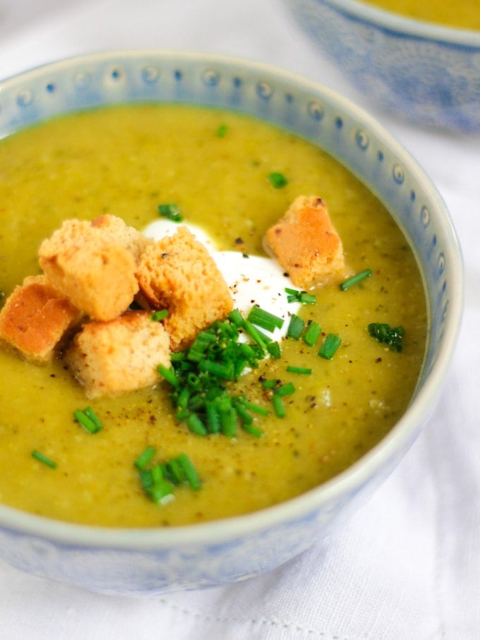 Side angle photo of a blue bowl of courgette soup recipe topped with creme fraiche, bread croutons and chives on white background.