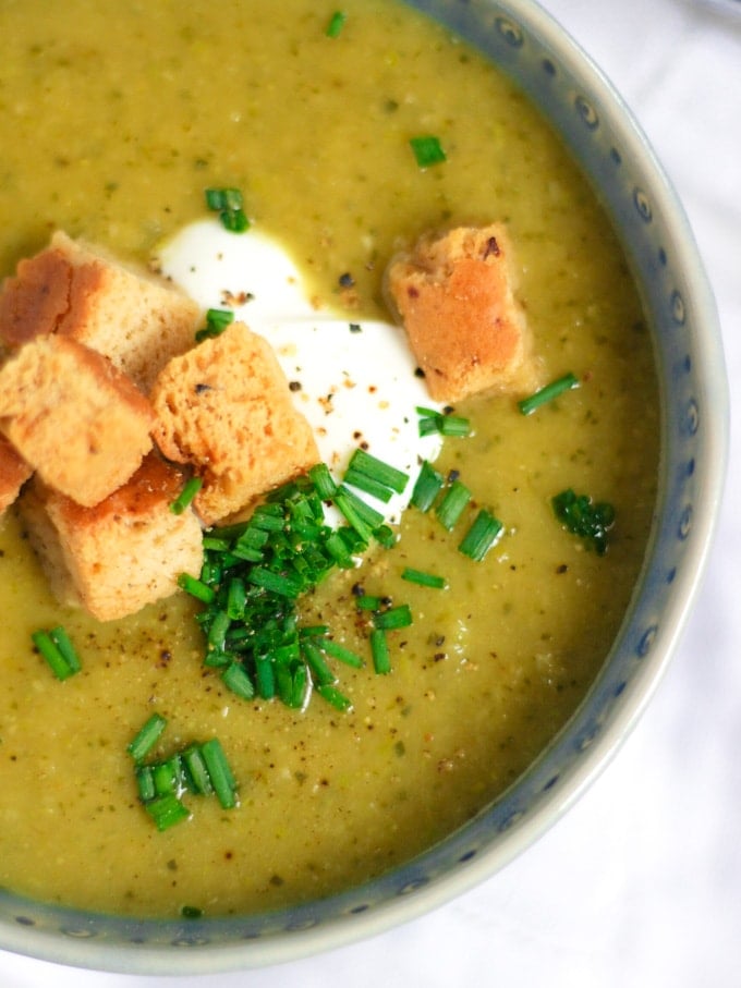 Overhead angle photo of a blue bowl of courgette soup recipe topped with creme fraiche, bread croutons and chives on white background.