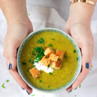Overhead angle photo of a blue bowl of courgette soup recipe being held in hands topped with creme fraiche, bread croutons and chives on white background.