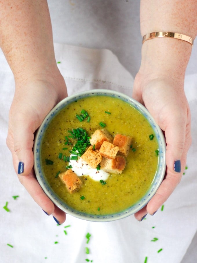 Overhead angle photo of a blue bowl of courgette soup recipe being held in hands topped with creme fraiche, bread croutons and chives on white background.