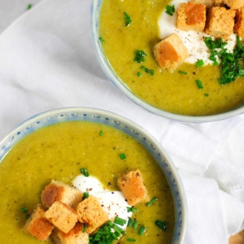 Overhead angle photo of a blue bowl of courgette soup recipe topped with creme fraiche, bread croutons and chives on white background.