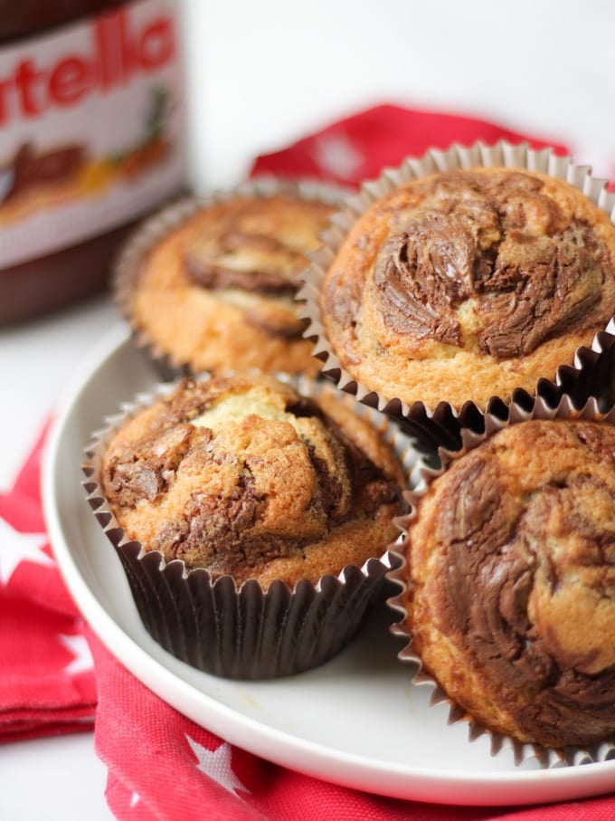 A pile of Nutella muffins in brown cupcake cases on a white plate on a red cloth with white stars and a jar of Nutella in the background.