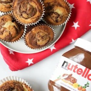 Overhead photo of a pile of Nutella muffins on a plate with a red cloth with white stars under it, one opened muffin and a jar of Nutella