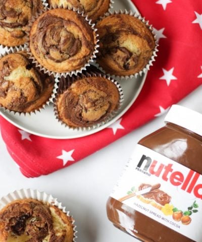 Overhead photo of a pile of Nutella muffins on a plate with a red cloth with white stars under it, one opened muffin and a jar of Nutella