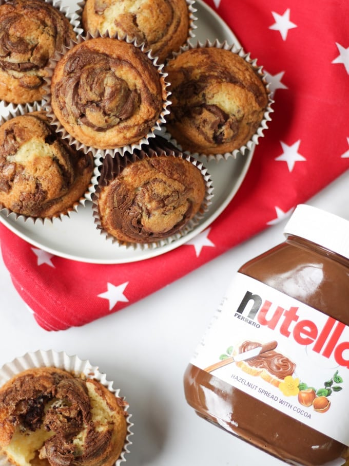 Overhead photo of a pile of Nutella muffins on a plate with a red cloth with white stars under it, one opened muffin and a jar of Nutella