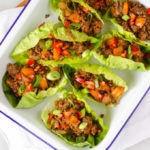 Cooked pork mince in lettuce leaves over head view in a white enamelware dish on white napkin, wooden board and white marble background.