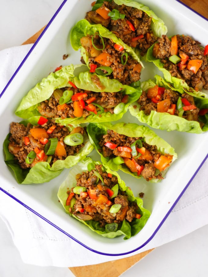 Cooked pork mince in lettuce leaves over head view in a white enamelware dish on white napkin, wooden board and white marble background.
