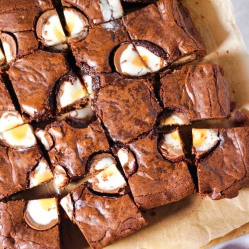 Creme Egg Brownie squares on baking paper on a white background with creme eggs in foil on the side