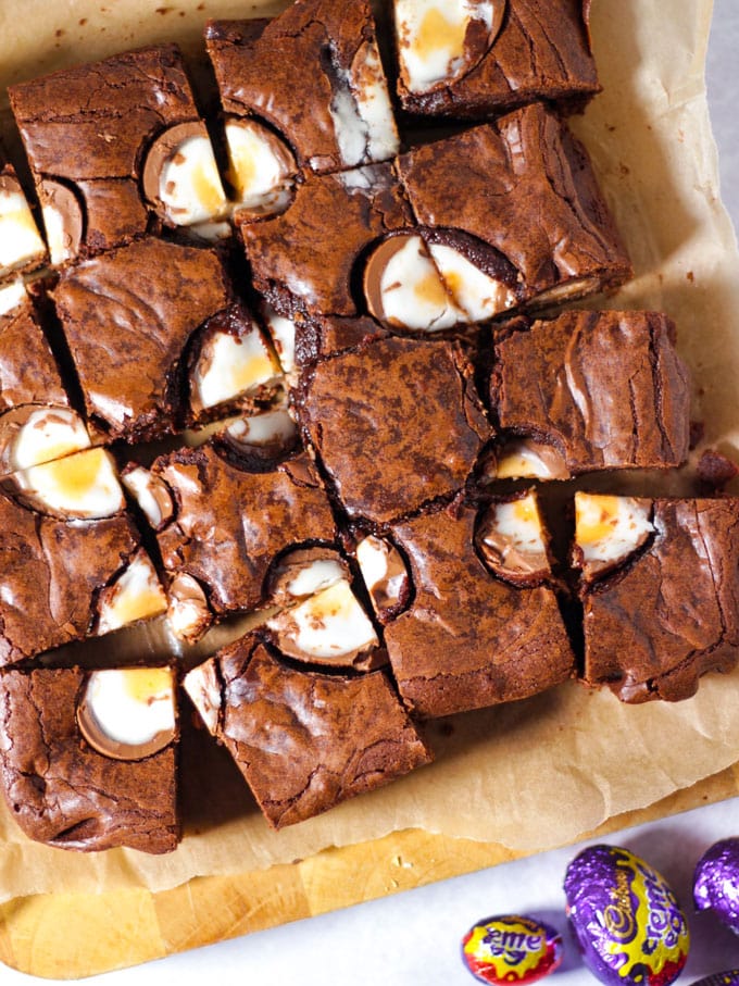 Creme Egg Brownies, cut into squares on a chopping board with foil wrapped creme eggs on the board and topped with cut chocolate creme eggs.