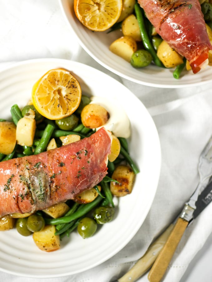 Chicken wrapped in bacon on white plates with green beans, olives and potatoes