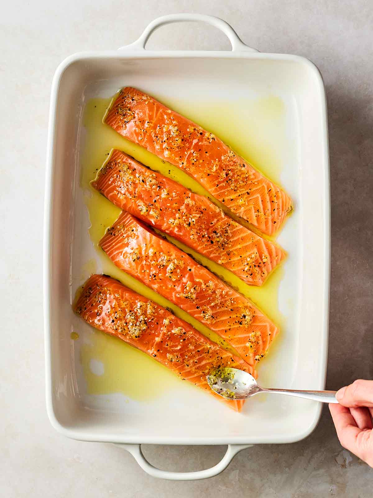 An oven dish with four pieces of raw salmon, with a dressing being put over the top with a spoon.