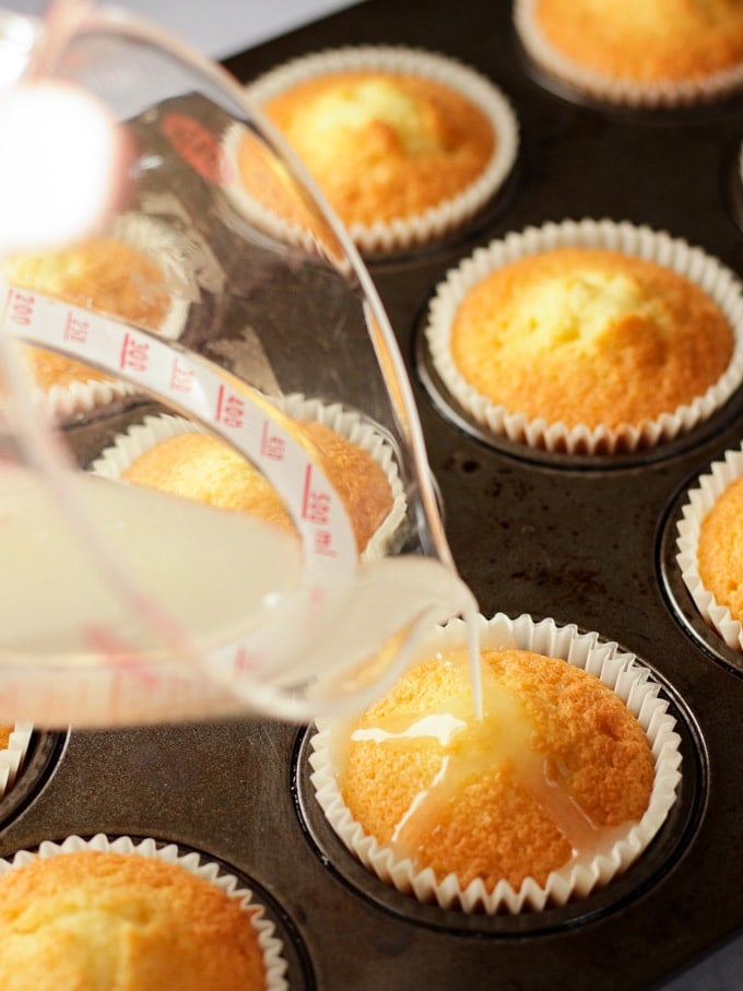 Baked cupcakes having lemon drizzle poured over them for step 3 in the recipe for Lemon Cupcakes.