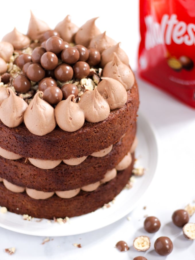 Malteser cake pilled with chocolate frosting and malt balls on a white plate