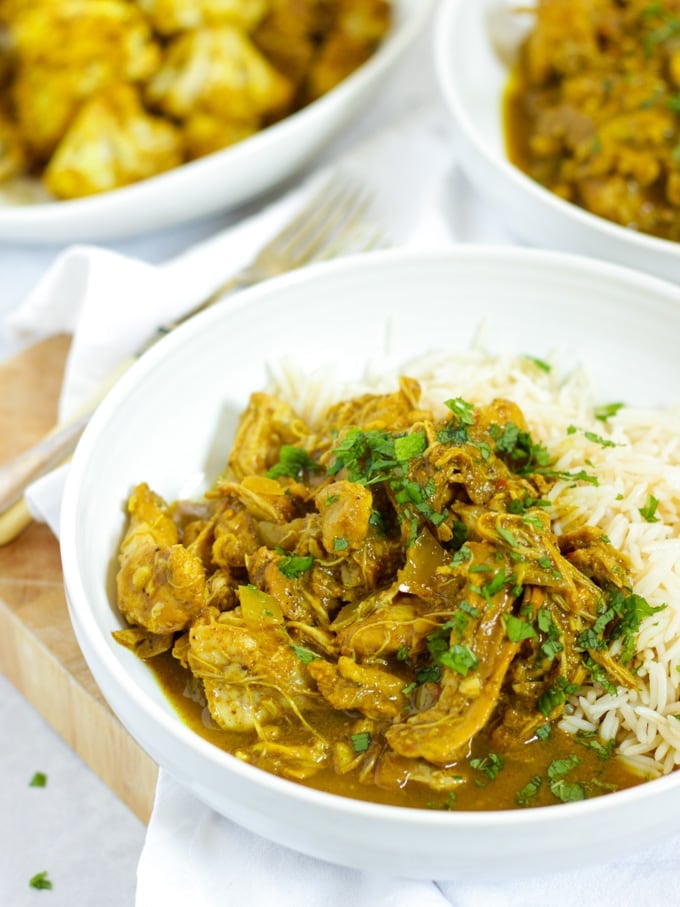 Bowl of slow cooker chicken curry with rice sprinkled with mint and with spiced roasted cauliflower in the background.