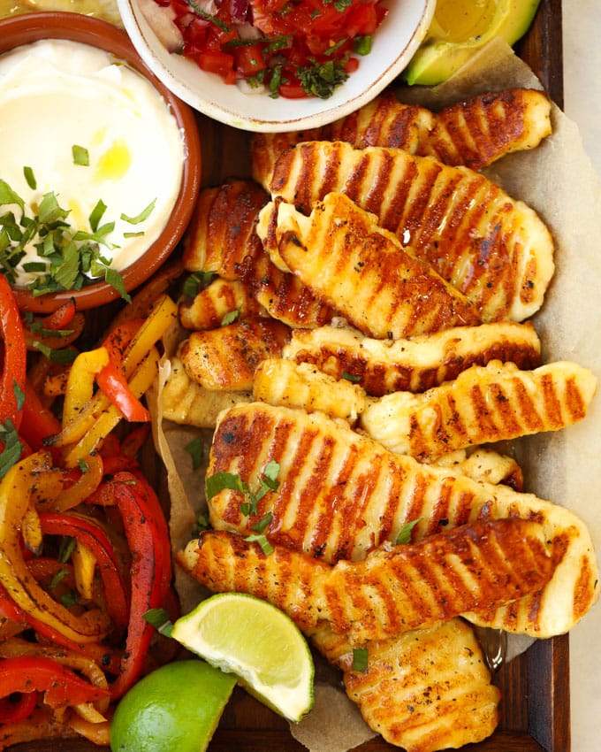 Grilled halloumi with griddle lines on, on a tray with vegetables, lime, sour cream and wraps for the recipe Halloumi Fajitas.