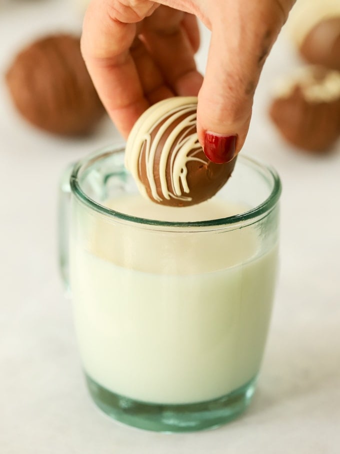 Mug of hot milk making cocoa with chocolate bomb about to be put in by a hand.