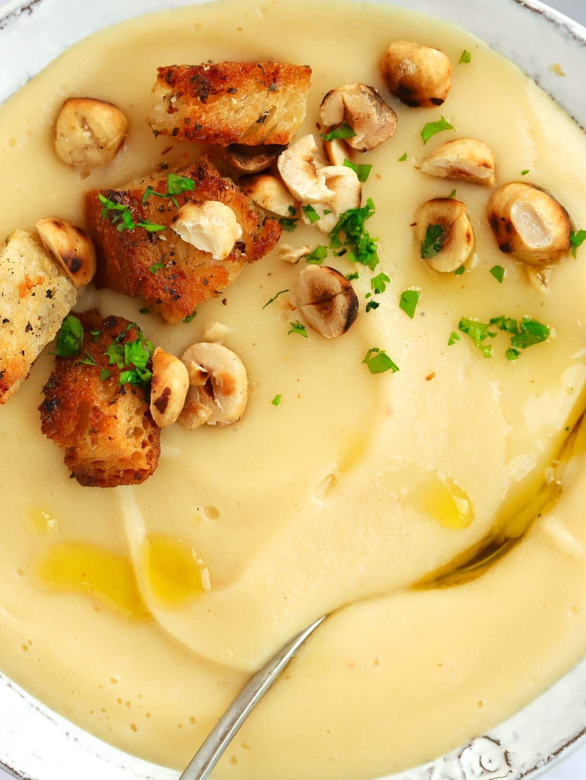 A close up of a creamy parsnip soup with croutons and hazelnuts on top, with a spoon ready to serve.