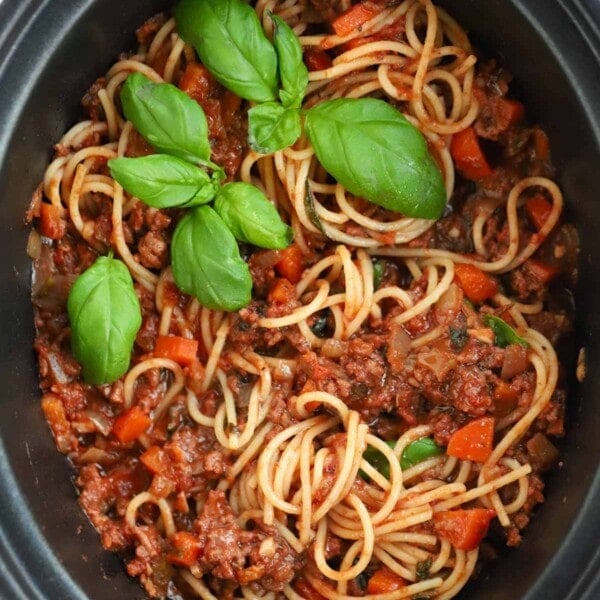 Slow cooker bolognese sauce in a slow cooker pan, mixed with cooked spaghetti and topped with basil.