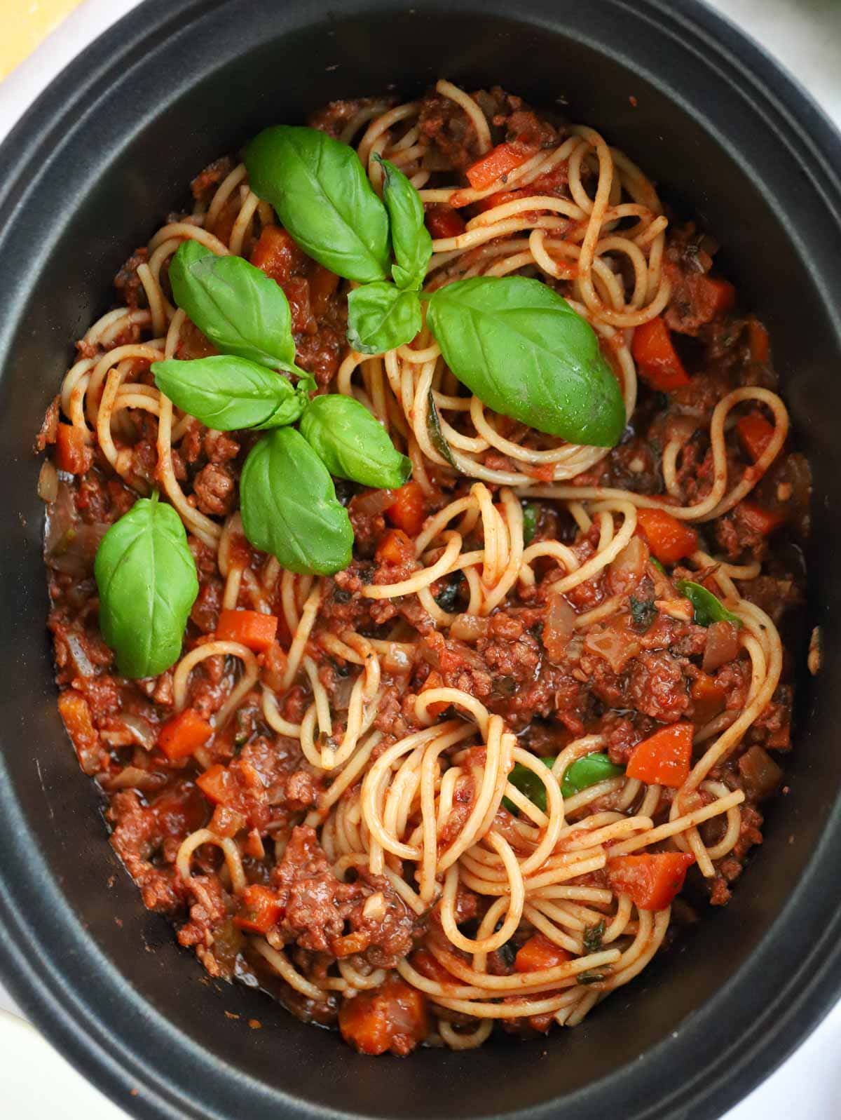 Slow cooker bolognese sauce in a slow cooker pan, mixed with cooked spaghetti and topped with basil. 